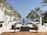 A luxurious outdoor space featuring an infinity pool flanked by tall palm trees. Modern, stylish furniture is arranged symmetrically around the pool, with white buildings aligning the sides. The setting suggests a serene and upscale environment, with clear skies and a distant view of the horizon.