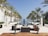 A luxurious outdoor space featuring an infinity pool flanked by tall palm trees. Modern, stylish furniture is arranged symmetrically around the pool, with white buildings aligning the sides. The setting suggests a serene and upscale environment, with clear skies and a distant view of the horizon.