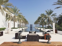 A luxurious outdoor space featuring an infinity pool flanked by tall palm trees. Modern, stylish furniture is arranged symmetrically around the pool, with white buildings aligning the sides. The setting suggests a serene and upscale environment, with clear skies and a distant view of the horizon.