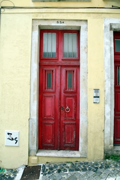 A rustic red double door set in a yellow wall, featuring worn wood panels and metal bars over frosted glass windows. The door has a prominent knocker ring, with a stone frame and electronic keypad on the right side. The ground is paved with uneven cobblestones, showing some grass between them.