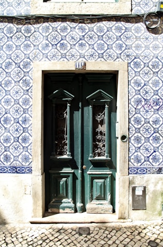 A set of double doors painted dark green, featuring intricate wrought iron designs in the windows, surrounded by a frame of weathered stone. The building facade is decorated with traditional blue and white Portuguese tiles with detailed floral patterns. A series of numbers above the door and a cobblestone pavement are visible in the foreground.