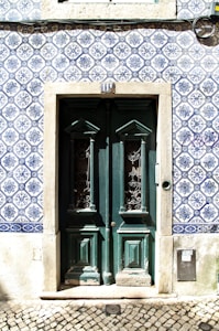 A set of double doors painted dark green, featuring intricate wrought iron designs in the windows, surrounded by a frame of weathered stone. The building facade is decorated with traditional blue and white Portuguese tiles with detailed floral patterns. A series of numbers above the door and a cobblestone pavement are visible in the foreground.