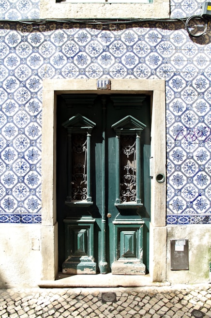 A set of double doors painted dark green, featuring intricate wrought iron designs in the windows, surrounded by a frame of weathered stone. The building facade is decorated with traditional blue and white Portuguese tiles with detailed floral patterns. A series of numbers above the door and a cobblestone pavement are visible in the foreground.