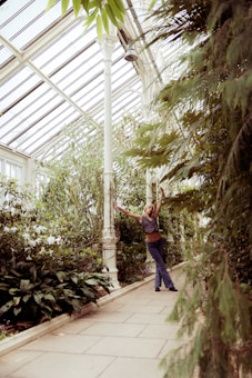 A person stands joyfully in a greenhouse surrounded by lush greenery. Tall plants and foliage create a vibrant atmosphere, with a pathway lined by a variety of plants. The glass-paneled ceiling lets in natural light, enhancing the verdant setting.