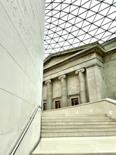 The image features a modern architectural interior combining classical and contemporary elements. A staircase in the foreground leads up to a facade with tall classical columns. The ceiling is a geometric patterned glass roof, allowing natural light to fill the space. Visible inscriptions run vertically along a wall on the left.