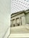 The image features a modern architectural interior combining classical and contemporary elements. A staircase in the foreground leads up to a facade with tall classical columns. The ceiling is a geometric patterned glass roof, allowing natural light to fill the space. Visible inscriptions run vertically along a wall on the left.