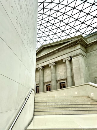 The image features a modern architectural interior combining classical and contemporary elements. A staircase in the foreground leads up to a facade with tall classical columns. The ceiling is a geometric patterned glass roof, allowing natural light to fill the space. Visible inscriptions run vertically along a wall on the left.