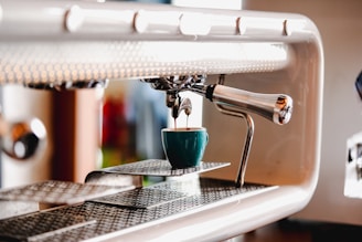 A modern espresso machine pours coffee into a small, turquoise cup placed on a metallic drip tray. The machine's chrome accents and sleek design suggest a high-end model. Soft lighting enhances the coffee-making process.