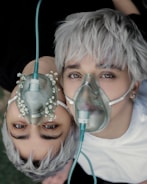 Two individuals with platinum blonde hair are lying close together, each wearing an oxygen mask. The masks are connected to a tube, and floral decorations are present on one mask, adding an artistic touch.