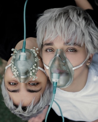 Two individuals with platinum blonde hair are lying close together, each wearing an oxygen mask. The masks are connected to a tube, and floral decorations are present on one mask, adding an artistic touch.