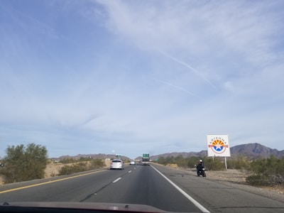 A highway stretches into the distance with vehicles in motion, surrounded by a desert landscape. A large sign with the Arizona state logo is visible on the right, and mountains are visible in the background under a clear sky.