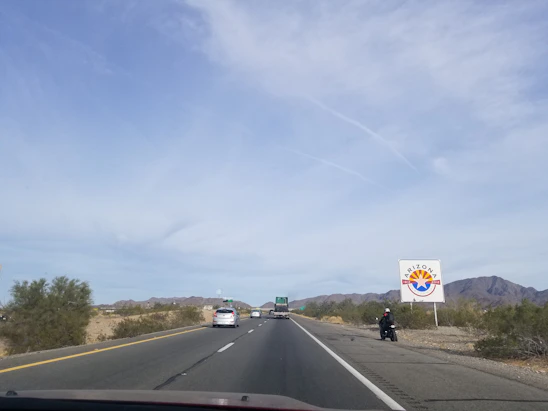 A highway stretches into the distance with vehicles in motion, surrounded by a desert landscape. A large sign with the Arizona state logo is visible on the right, and mountains are visible in the background under a clear sky.