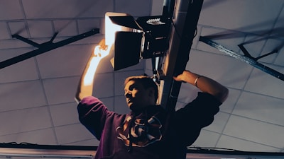A person is adjusting a stage light fixture mounted on a ceiling. The lighting creates dramatic shadows, highlighting the person's focused expression as they work. The setting appears to be industrial or stage-like with visible structural elements.