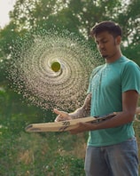 A young man in a teal shirt and jeans is skillfully balancing a cricket bat with a spinning tennis ball above it. The ball is wet, creating a spiral pattern of water droplets in mid-air. The background is lush and green with blurred foliage.