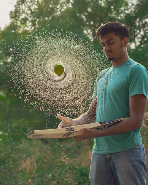 A young man in a teal shirt and jeans is skillfully balancing a cricket bat with a spinning tennis ball above it. The ball is wet, creating a spiral pattern of water droplets in mid-air. The background is lush and green with blurred foliage.