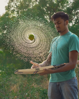 A young man in a teal shirt and jeans is skillfully balancing a cricket bat with a spinning tennis ball above it. The ball is wet, creating a spiral pattern of water droplets in mid-air. The background is lush and green with blurred foliage.