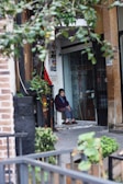 An elderly person sits on a white chair at the entrance of a building, surrounded by a mix of urban and natural elements. The scene includes leafy branches in the foreground and a variety of textures such as brick and concrete. The individual, dressed in a dark sweater and slippers, has a contemplative expression.