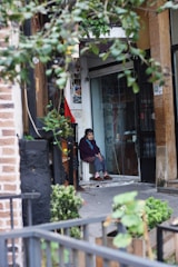 An elderly person sits on a white chair at the entrance of a building, surrounded by a mix of urban and natural elements. The scene includes leafy branches in the foreground and a variety of textures such as brick and concrete. The individual, dressed in a dark sweater and slippers, has a contemplative expression.