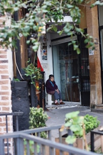 An elderly person sits on a white chair at the entrance of a building, surrounded by a mix of urban and natural elements. The scene includes leafy branches in the foreground and a variety of textures such as brick and concrete. The individual, dressed in a dark sweater and slippers, has a contemplative expression.