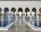 An architectural scene featuring ornate columns adorned with gold accents and intricate designs. The columns line a reflective water pool with a polished tile surface, leading toward a vast courtyard. The setting is tranquil, with only a few people walking in the distance.