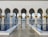 An architectural scene featuring ornate columns adorned with gold accents and intricate designs. The columns line a reflective water pool with a polished tile surface, leading toward a vast courtyard. The setting is tranquil, with only a few people walking in the distance.
