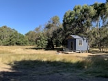 A small, modern tiny house on wheels is situated in a grassy field surrounded by tall trees. The structure is painted in a subdued grayish-blue color and features a window and a small outdoor deck with a table and chairs.