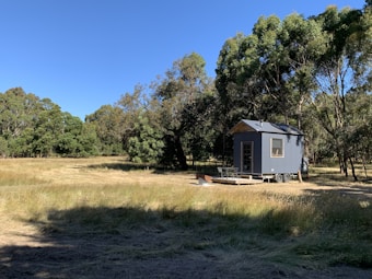 A small, modern tiny house on wheels is situated in a grassy field surrounded by tall trees. The structure is painted in a subdued grayish-blue color and features a window and a small outdoor deck with a table and chairs.