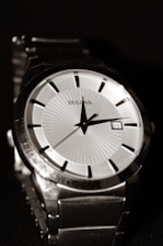 A close-up view of a luxury wristwatch featuring a sleek design with a metallic silver band, a white dial, and black hour markers. The hands of the watch point to the hour and minute, with a small date window showing the number 23. The watch brand name is visible on the dial.