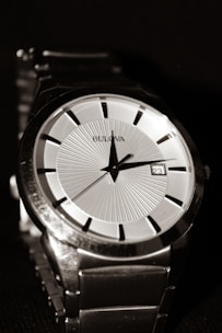 A close-up view of a luxury wristwatch featuring a sleek design with a metallic silver band, a white dial, and black hour markers. The hands of the watch point to the hour and minute, with a small date window showing the number 23. The watch brand name is visible on the dial.