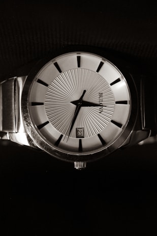 A classic wristwatch with a metallic band and a round face, featuring a detailed textured dial with black hour markers. The hands are sleek and the watch displays the date on the dial.
