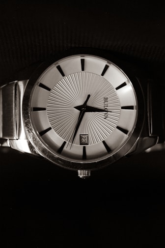 A classic wristwatch with a metallic band and a round face, featuring a detailed textured dial with black hour markers. The hands are sleek and the watch displays the date on the dial.