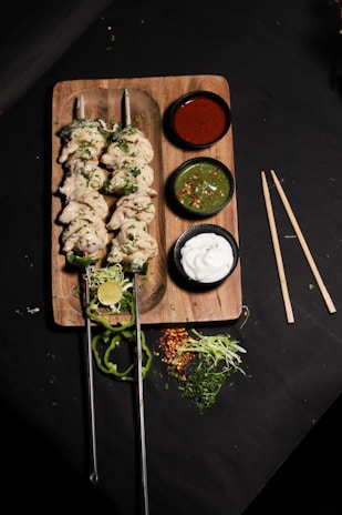 A wooden board holds two skewers of cooked chicken kabobs garnished with herbs. Accompanying the skewers are three small bowls containing sauces: a red sauce, a green chutney with chili flakes, and a white yogurt-based dip. Freshly cut lime, slices of green pepper, shredded cabbage, and other greens are scattered around the board, adding a fresh touch.