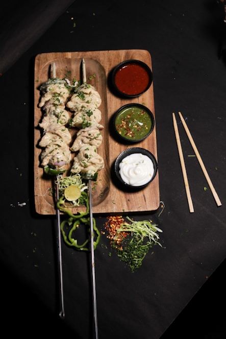 A wooden board holds two skewers of cooked chicken kabobs garnished with herbs. Accompanying the skewers are three small bowls containing sauces: a red sauce, a green chutney with chili flakes, and a white yogurt-based dip. Freshly cut lime, slices of green pepper, shredded cabbage, and other greens are scattered around the board, adding a fresh touch.