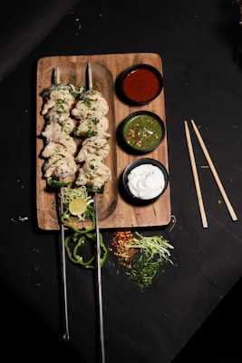 A wooden board holds two skewers of cooked chicken kabobs garnished with herbs. Accompanying the skewers are three small bowls containing sauces: a red sauce, a green chutney with chili flakes, and a white yogurt-based dip. Freshly cut lime, slices of green pepper, shredded cabbage, and other greens are scattered around the board, adding a fresh touch.
