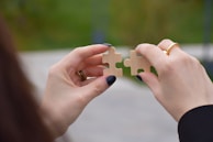A close-up view of two hands holding puzzle pieces that are being fit together. The nails are painted dark, and the background is softly blurred with green and gray tones.