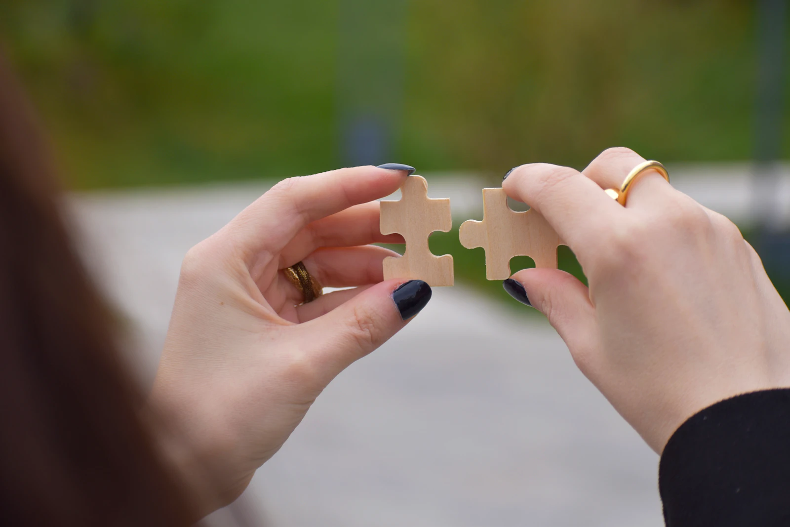 Two hands holding wooden puzzle pieces together against a blurred outdoor background