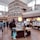 A modern library with tall shelves filled with books stretches up to the ceiling. Customers browse through magazines and books on display tables. Large windows allow natural light to fill the spacious, two-story area featuring a wooden floor and contemporary design.