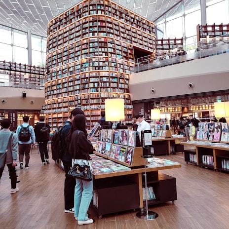 A modern library with tall shelves filled with books stretches up to the ceiling. Customers browse through magazines and books on display tables. Large windows allow natural light to fill the spacious, two-story area featuring a wooden floor and contemporary design.