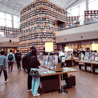 A modern library with tall shelves filled with books stretches up to the ceiling. Customers browse through magazines and books on display tables. Large windows allow natural light to fill the spacious, two-story area featuring a wooden floor and contemporary design.