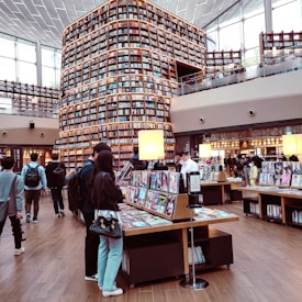 A modern library with tall shelves filled with books stretches up to the ceiling. Customers browse through magazines and books on display tables. Large windows allow natural light to fill the spacious, two-story area featuring a wooden floor and contemporary design.