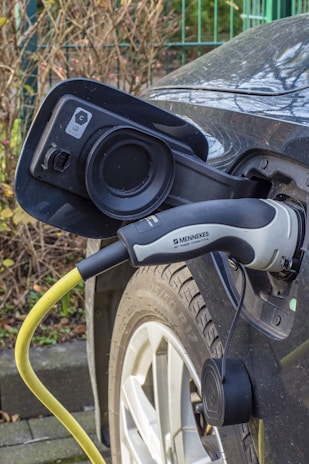 A close-up view of an electric vehicle charging port with a charging cable attached. The cable is yellow and connected to a car, whose wheel and part of the body are visible. The brand name 'MENNEKES' is printed on the charging handle. Background shows some greenery and a metal fence.