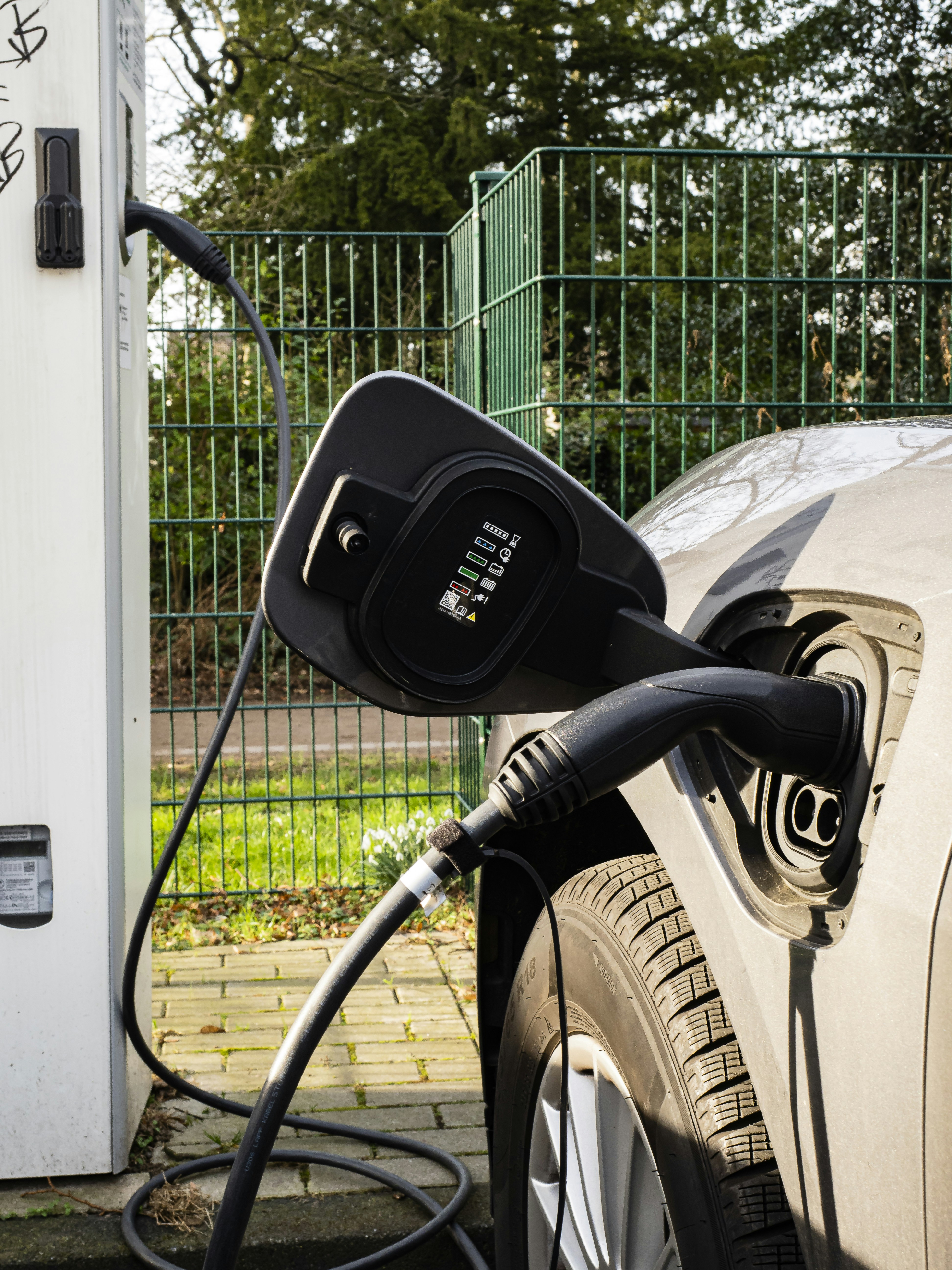 An electric vehicle is plugged into a charging station. The image captures a close-up view of the charging port of the car and the connected charging cable. The vehicle is parked on a concrete path near a metal fence, with greenery visible in the background.
