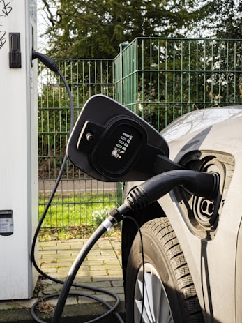 An electric vehicle is plugged into a charging station. The image captures a close-up view of the charging port of the car and the connected charging cable. The vehicle is parked on a concrete path near a metal fence, with greenery visible in the background.