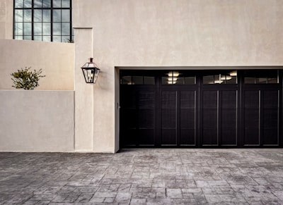 A modern garage door with a dark finish set against a light-colored stucco wall. A traditional-style lamp is mounted on the wall, and a small potted plant is visible on a ledge. The driveway is paved with patterned stone.