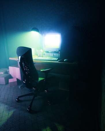 A dimly lit room features a vintage computer setup on a wooden desk with a CRT monitor emitting a bright glow. The scene includes an ergonomic office chair positioned in front of the desk. Several cardboard boxes are stacked on the carpeted floor nearby, and a lamp provides soft greenish lighting, creating a serene yet mysterious ambiance.