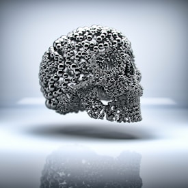 A detailed representation of a skull created from numerous small, shiny metallic spheres, giving it a futuristic and abstract appearance. The skull is suspended in mid-air against a soft, blurred background.
