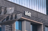 A modern building facade features a sign with 'Wolt' written in a white, cursive font against dark, tiled exterior walls. The structure has large windows with reflections, and there is a metallic overhang on the left side.