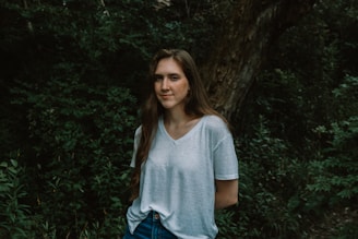 A young woman with long brown hair stands in front of dense foliage and a tree trunk. She is wearing a white T-shirt and blue jeans, with her hands behind her back. The setting is natural, with lush greenery surrounding her.