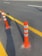 Several orange and white traffic delineator posts are arranged alongside freshly painted yellow lines on an asphalt road. The long shadows suggest it is either early morning or late afternoon.