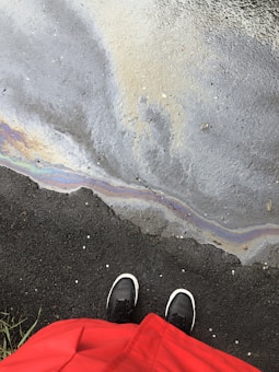 A person in a red coat stands on wet asphalt next to a puddle with an iridescent oily sheen. Black and white shoes are visible, and there are some small patches of grass at the border of the pavement.
